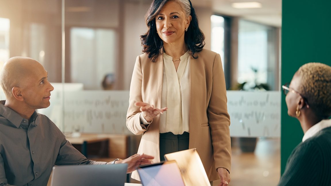Mujer de pie dirigiendo una conversación con dos colegas sentados frente a computadoras — estilos de liderazgo